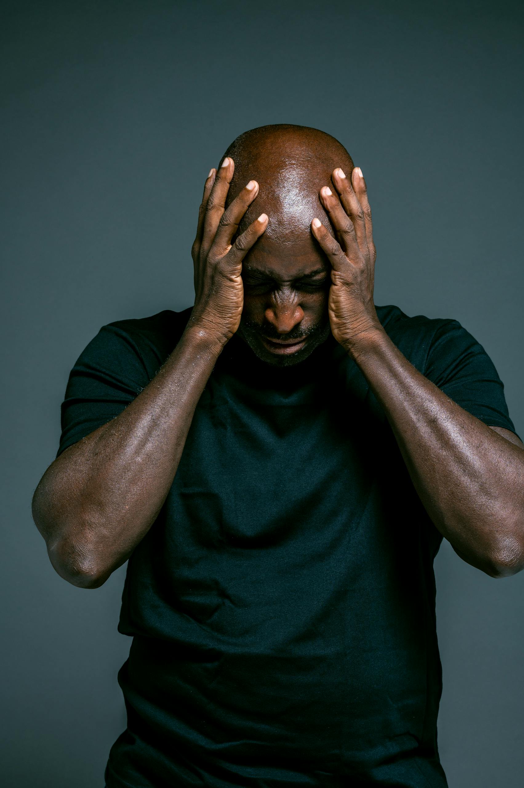 Bald African American man in stress with hands on head against dark background.